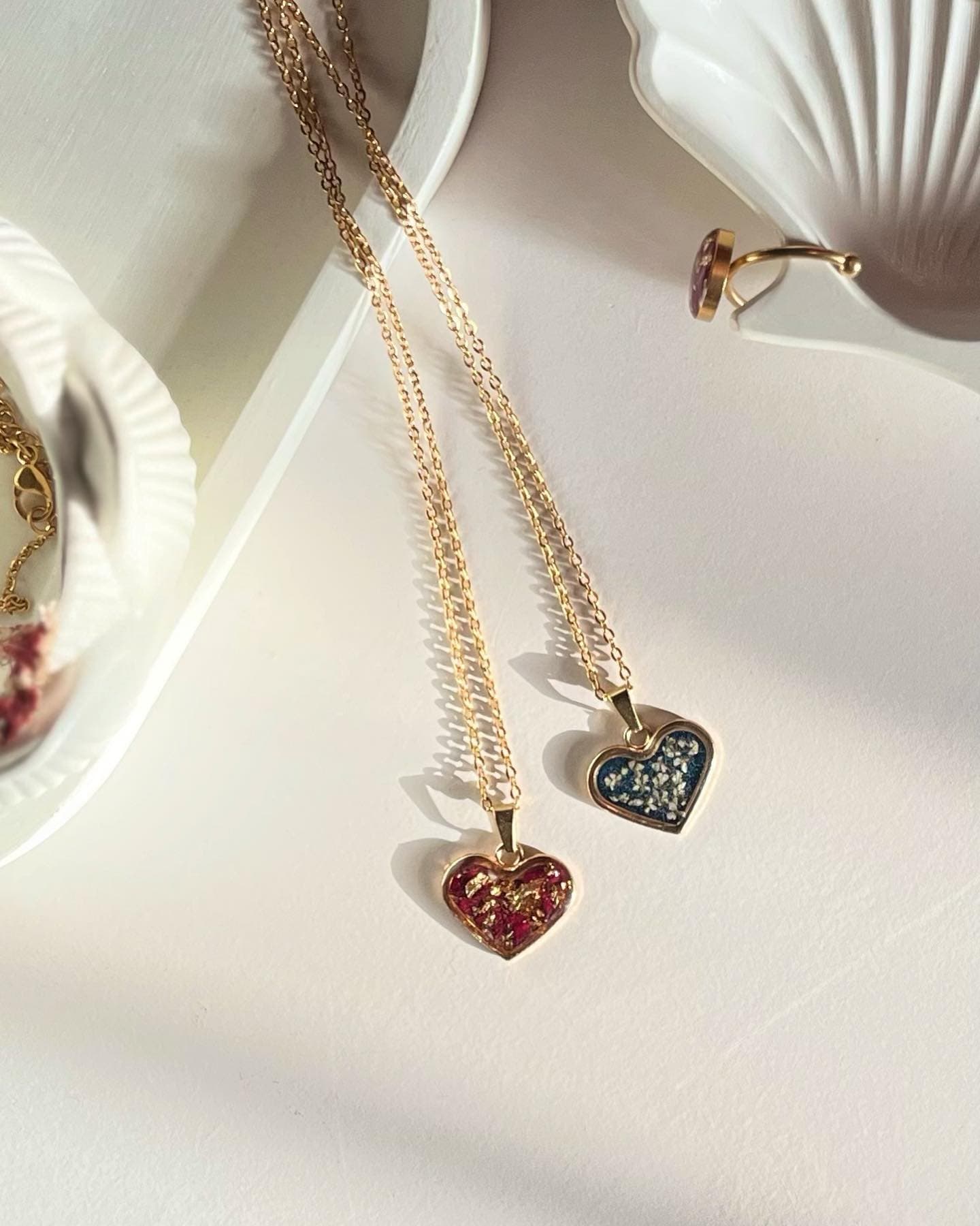 Two small gold heart pendants on fine chains, one deep red with gold leaf inside and one navy blue with pressed white flowers